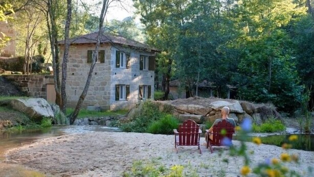 A stone house by a river with a wooden chair and a bench.