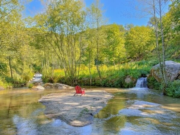 Un entorno exterior tranquilo con una pequeña cascada, una silla roja y abundante vegetación.