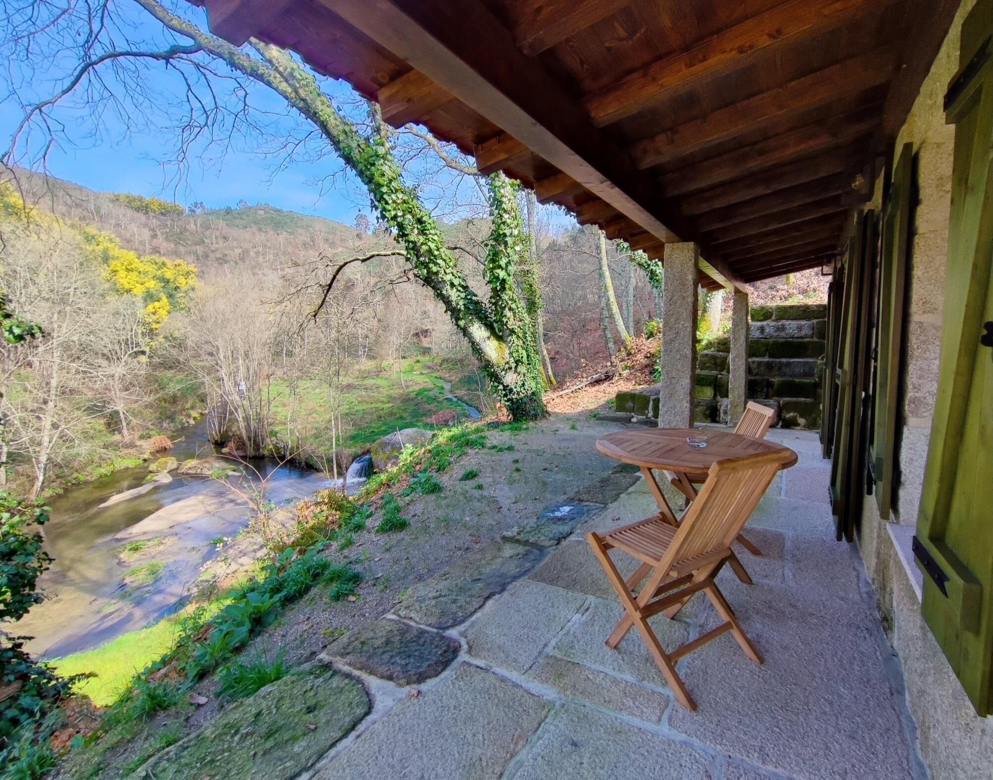 Un patio de piedra con una mesa y sillas de madera, con vistas a un arroyo y una ladera boscosa.