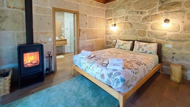 Quarto com parede de pedra, teto de madeira, cama com roupa de cama floral e lareira a lenha.