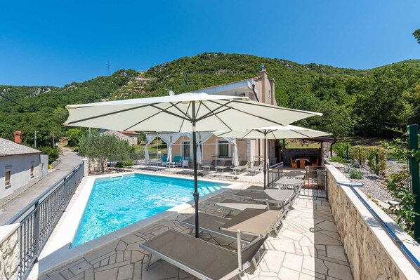 Zona de piscina con sillas de descanso y sombrillas, rodeada por un muro de piedra y vistas a una colina.