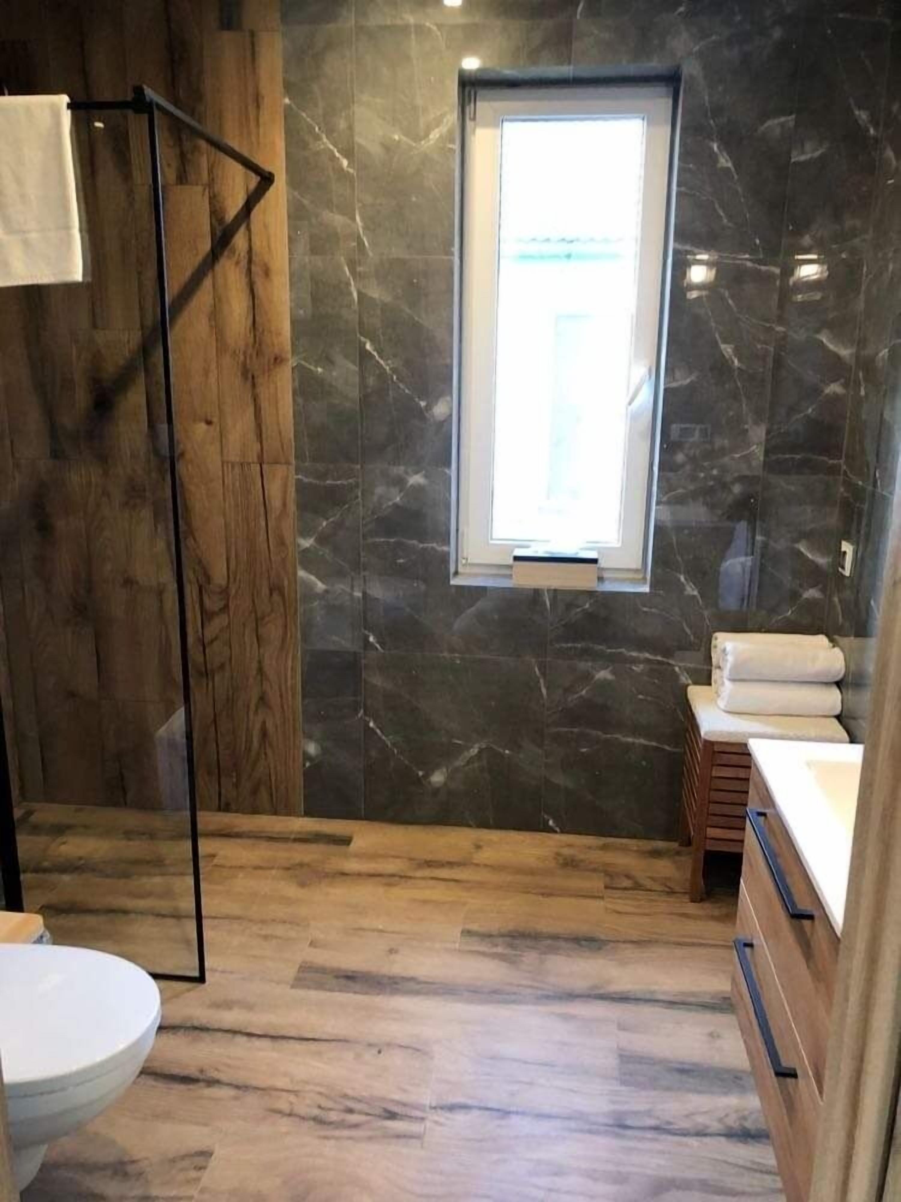 A bathroom with a wooden floor, a marble wall, a window, a toilet, and a wooden vanity with a sink.