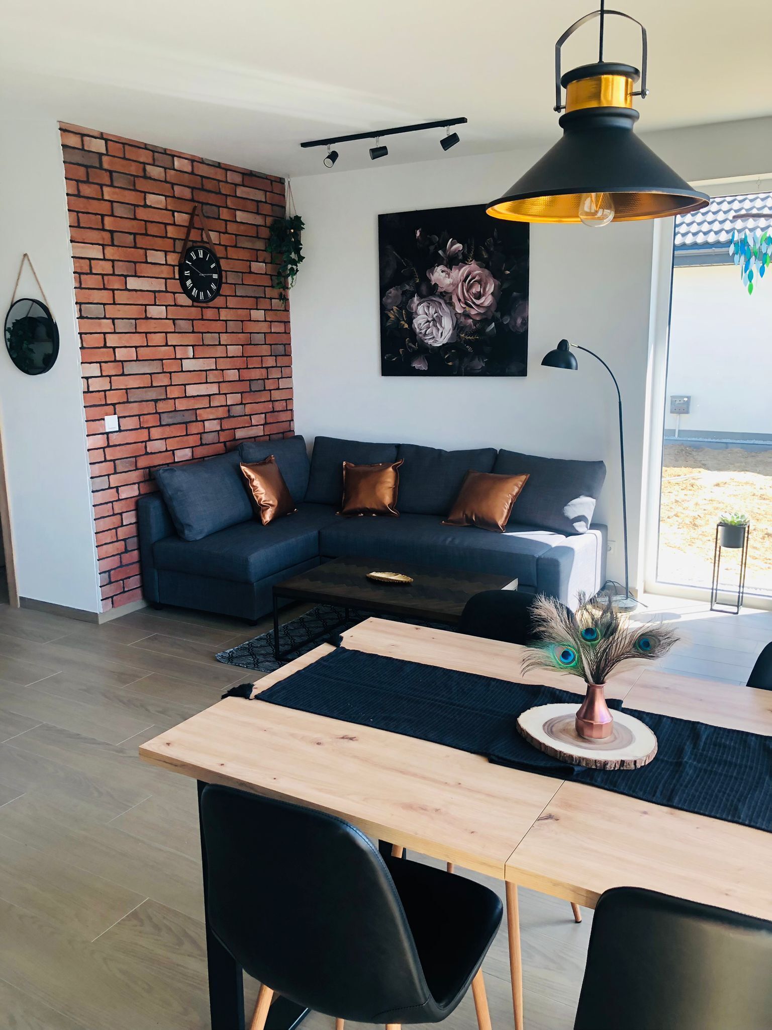 Una sala moderna con una pared de acento de ladrillo, un sofá azul oscuro, una mesa de comedor de madera y una decoración con plumas de pavo real.