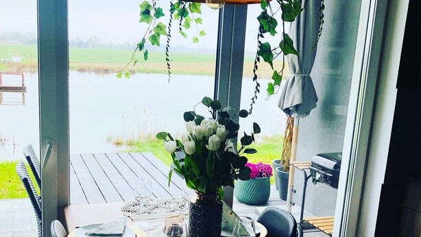 Ein Essbereich mit Tisch für zwei Personen, einem Blumentopf und Blick auf ein Gewässer durch große Fenster.