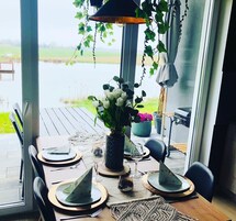 Un área de comedor con una mesa dispuesta para dos personas, una planta en maceta y vistas a una masa de agua a través de amplias ventanas.