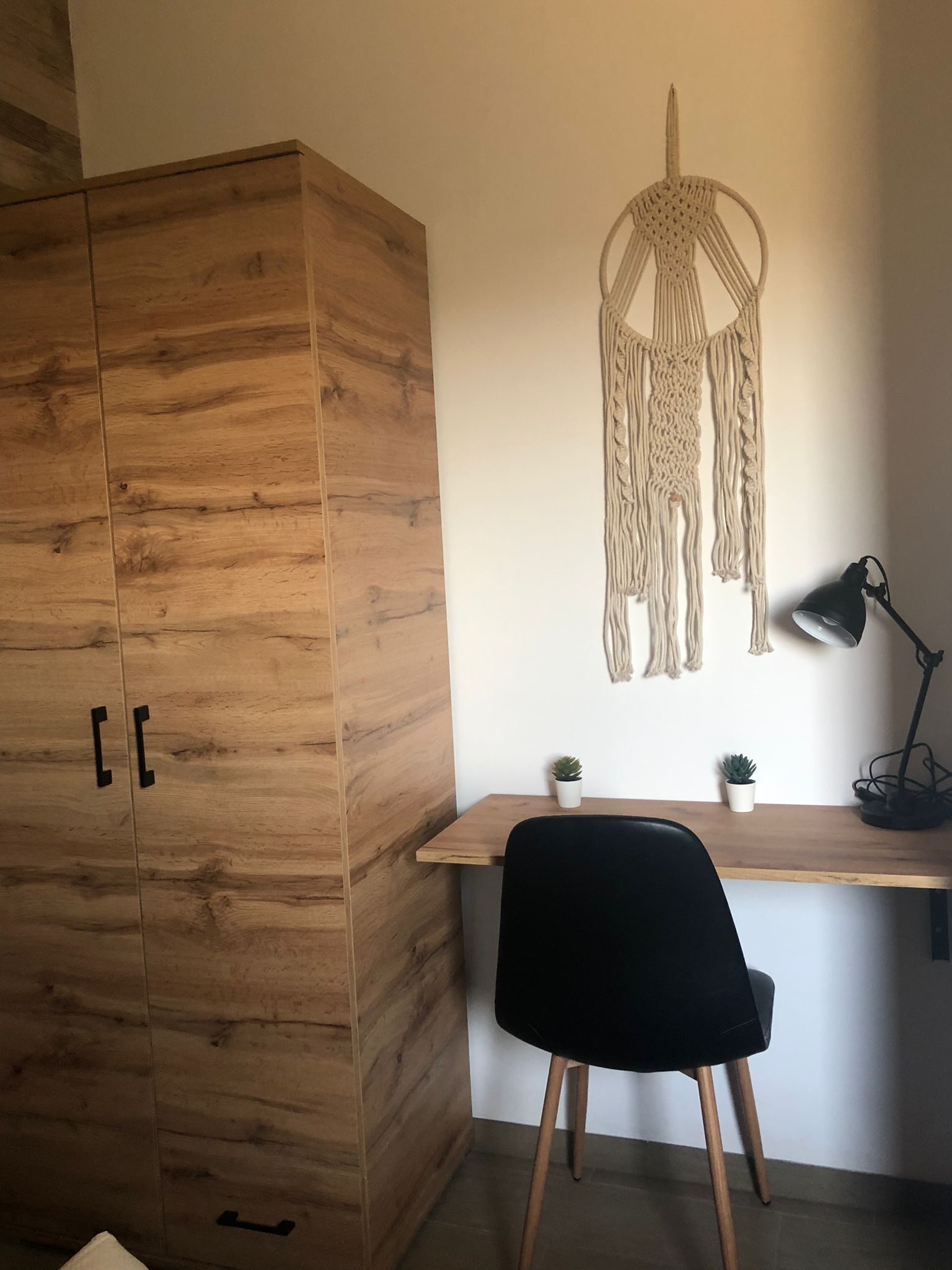 Un armario de madera con dos tiradores, un escritorio con una silla negra, un colgador de macramé en la pared y dos pequeñas macetas con plantas.