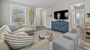 A living room with a beige sofa, striped cushions, a round coffee table, and a blue cabinet with a TV mounted above it. - 5 Min Stroll to Beach in PCB: Gm Rm+Put Grn+Yard! (Panama City Beach)