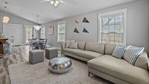A spacious living room with a beige sofa, round ottomans, and a coffee table. There is a dining area with a table and chairs, and a ceiling fan. - 5 Min Stroll to Beach in PCB: Gm Rm+Put Grn+Yard! (Panama City Beach)