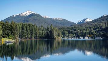Um lago tranquilo com reflexos de montanhas e árvores, rodeado por uma área arborizada.