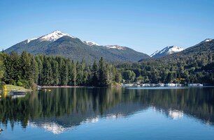 Um lago tranquilo com reflexos de montanhas e árvores, rodeado por uma área arborizada.
