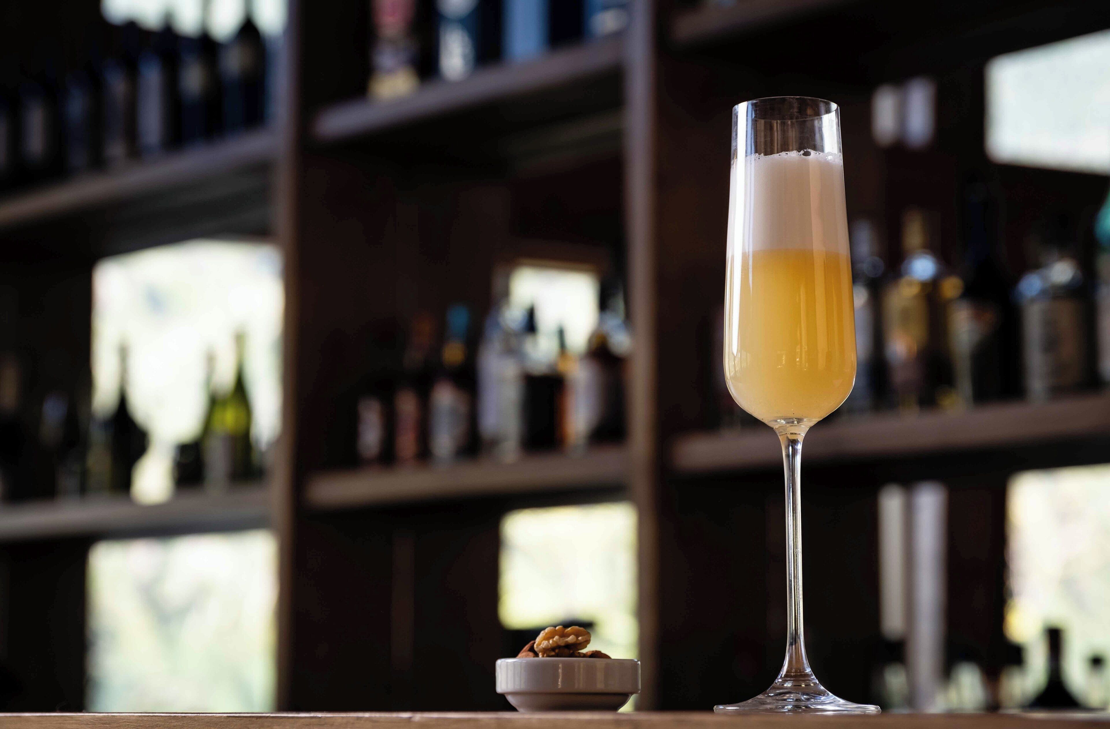 A tall glass of beer on a bar counter with a small bowl of nuts beside it.