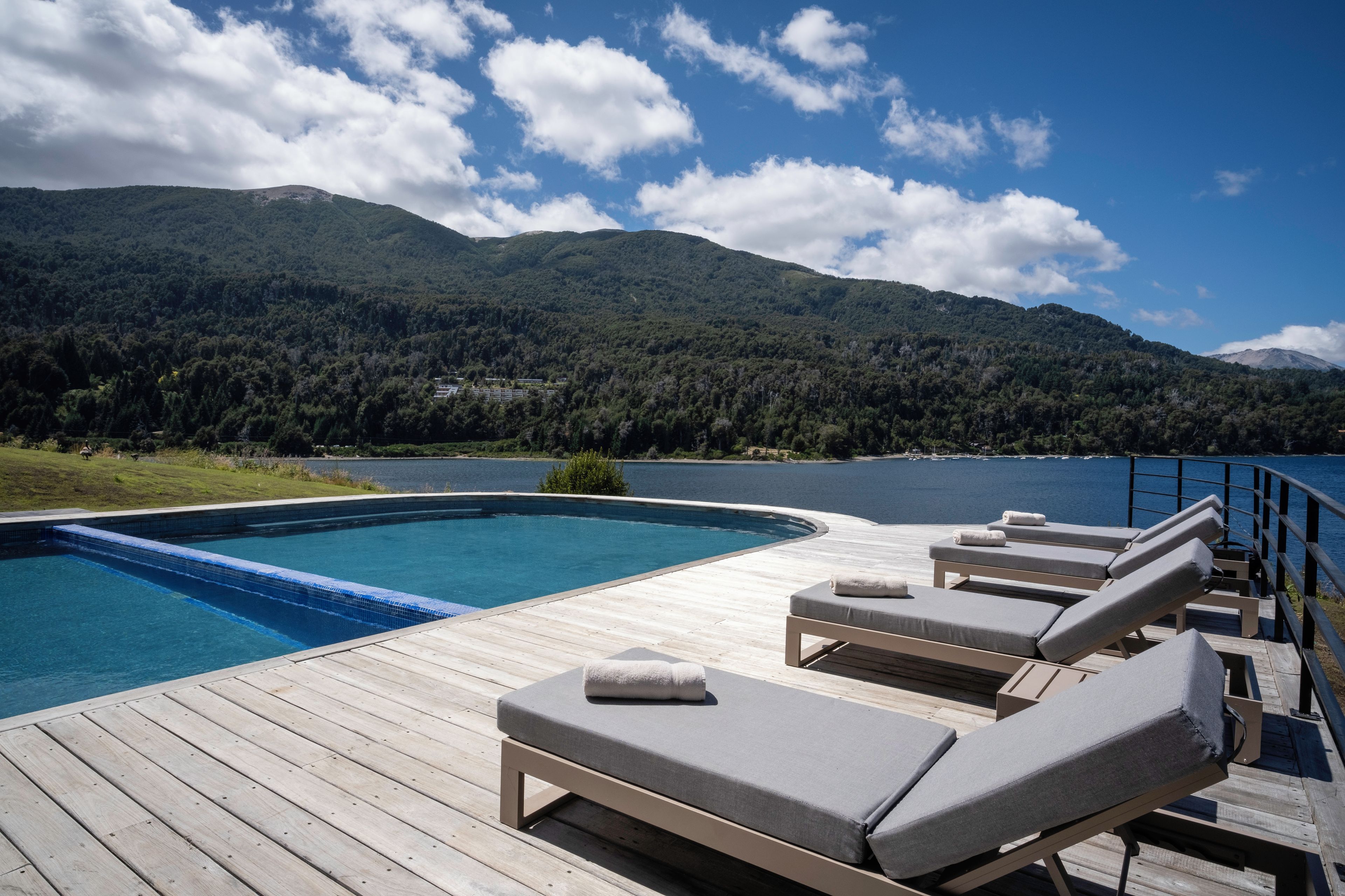 Terraza de madera con piscina y sillones de descanso con vista a un lago y montañas al fondo.
