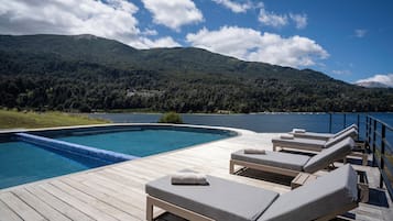 Terraza de madera con piscina y sillones de descanso con vista a un lago y montañas al fondo.
