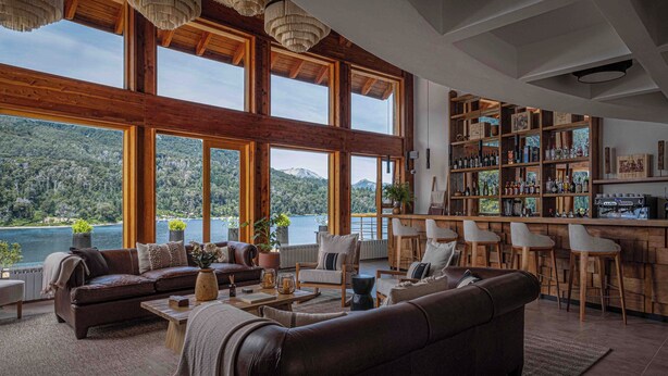 Amplia sala de estar con un ventanal que da a un lago y montañas, amoblada con sofás de cuero y un área de bar con estanterías de madera.