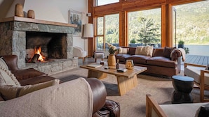A cozy living room with a stone fireplace, leather furniture, and large windows offering a view of the forest. - Puerto Manzano Boutique Hotel (Villa La Angostura)