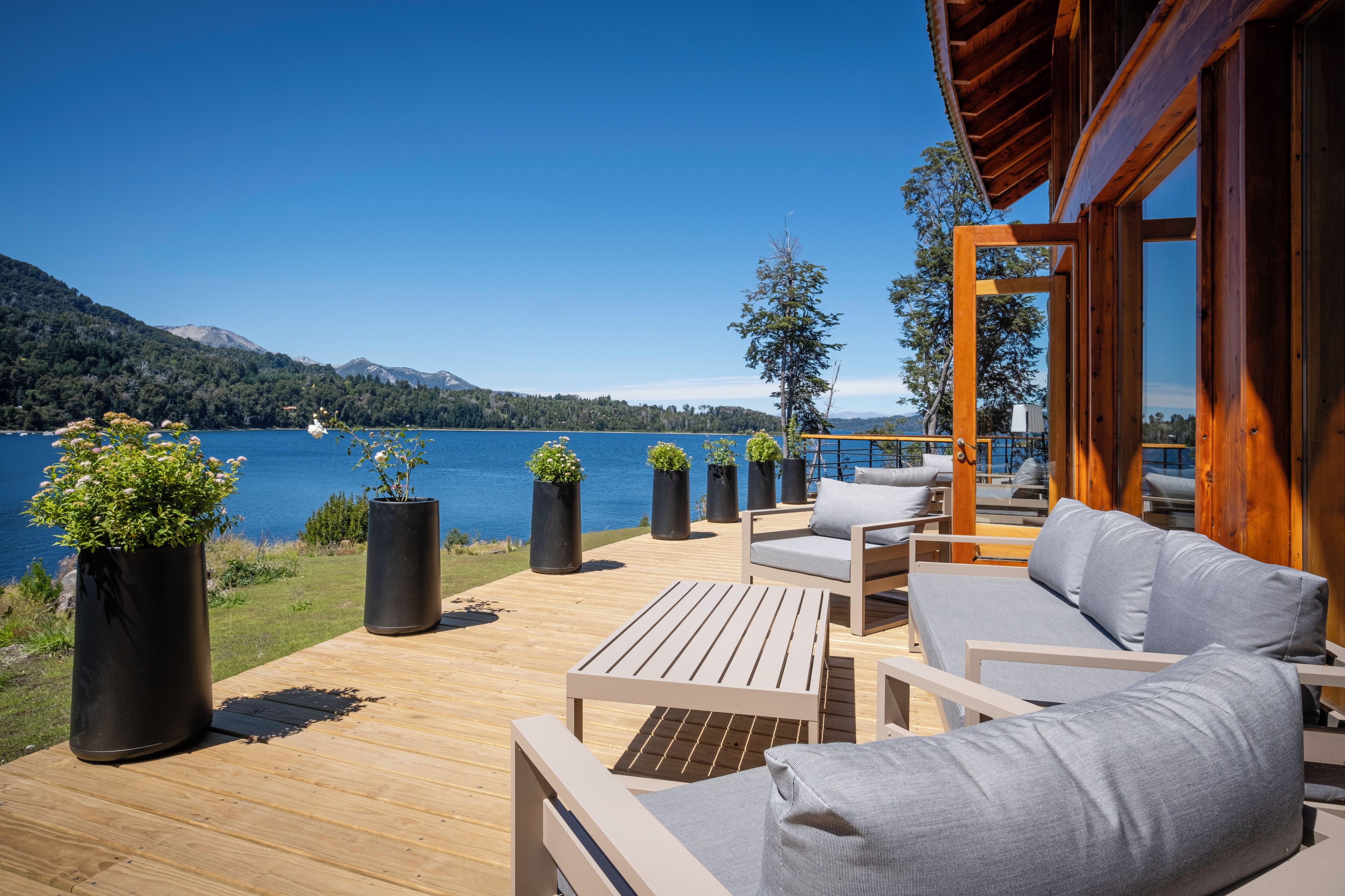 Terraza de madera con mobiliario exterior que da a un lago, rodeada de plantas en macetas.