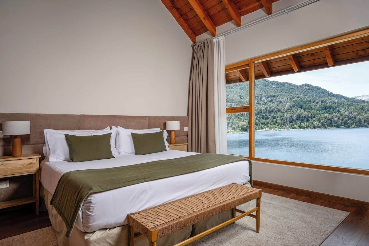 Une chambre avec une grande fenêtre, un lit avec un oreiller vert, un banc en bois et une table de chevet avec une lampe.