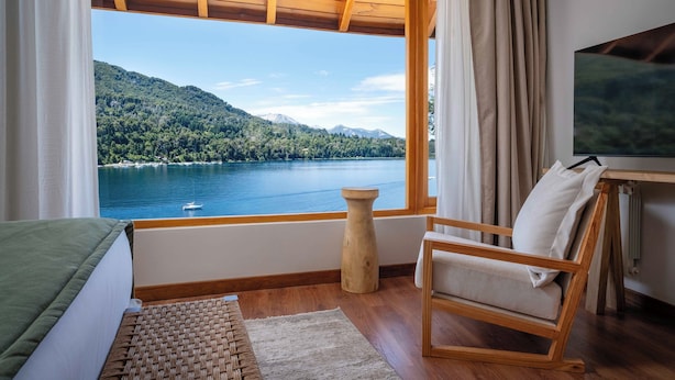 A room with a wooden floor, a bed with a green cover, a wooden chair, a small table with a TV, and a large window with a view of a lake and mountains. - Puerto Manzano Boutique Hotel (Villa La Angostura)