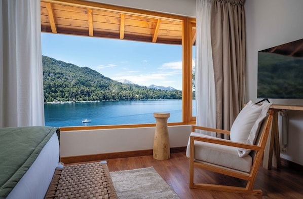 A room with a wooden floor, a bed with a green cover, a wooden chair, a small table with a TV, and a large window with a view of a lake and mountains. - Puerto Manzano Boutique Hotel (Villa La Angostura)