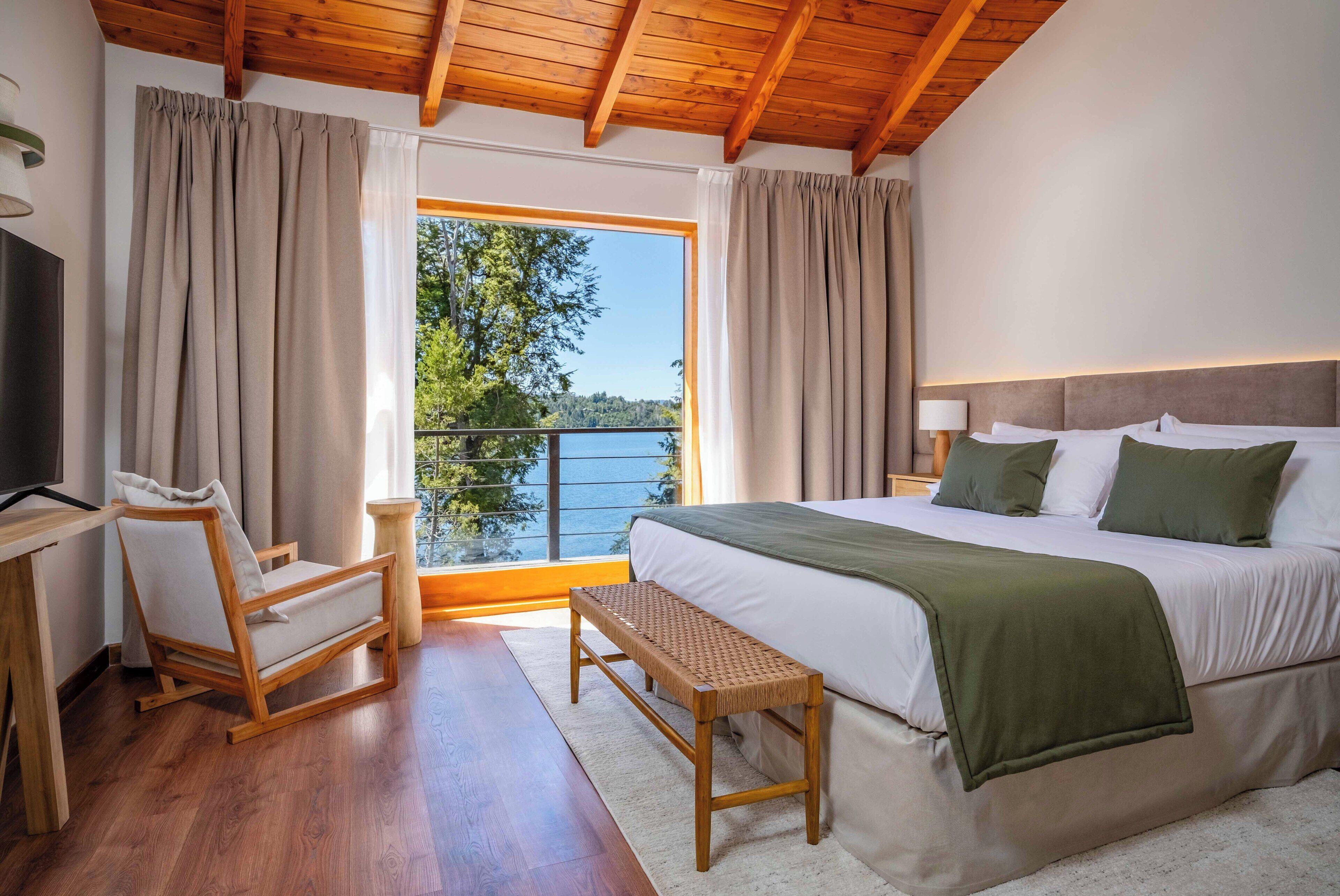 Un dormitorio con cama, un banco de madera, televisor y vista a un lago.