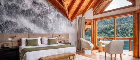 A bedroom with a large bed, a bench, a chair, and a view of a forest through large windows.