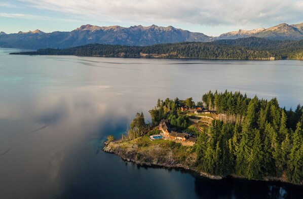 A lakeside property with a large house surrounded by dense forest, a swimming pool, and mountains in the background.