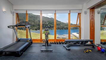 A gym with a treadmill, elliptical, bench, and dumbbells, overlooking a lake and mountains.