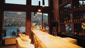 Zona de bar con techo de madera, amplias ventanas con vistas a un lago y barra llena de botellas.