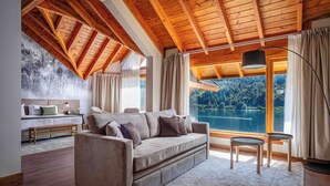 A cozy living room with a large window overlooking a lake, a wooden ceiling, and a sofa with cushions. - Puerto Manzano Boutique Hotel (Villa La Angostura)
