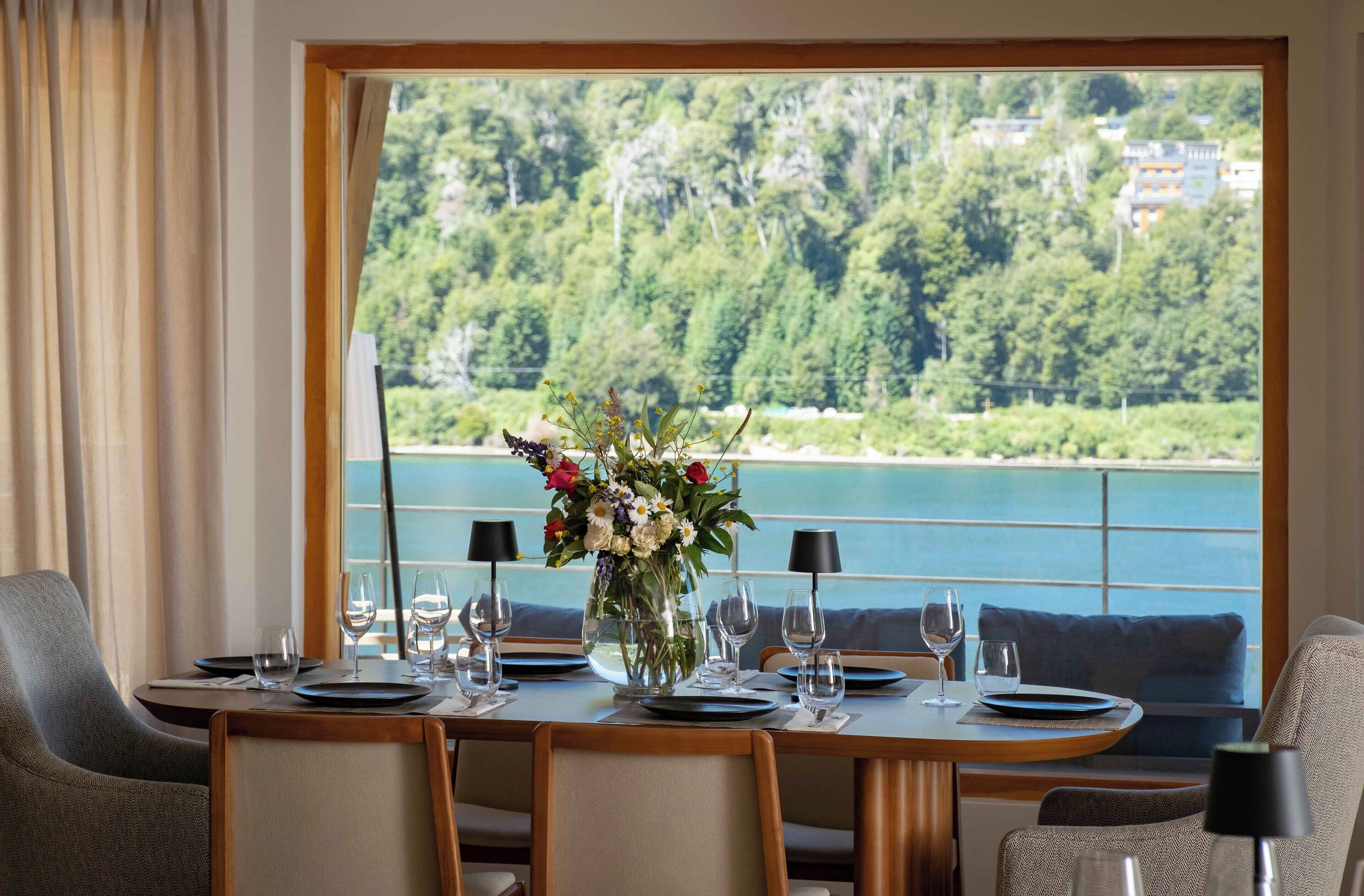 Uma mesa de jantar posta para quatro, com um vaso de flores, duas luminárias e vista para um lago e uma floresta.