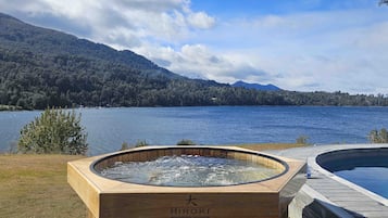 Uma banheira de madeira com uma etiqueta que diz "Hiroki Minipiscina Japonesa" à beira de um lago, com montanhas ao fundo.