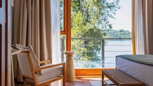 A room with a bed, a wooden chair, a small table, and a view of a lake through large windows. - Puerto Manzano Boutique Hotel (Villa La Angostura)