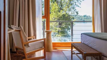 Una habitación con una cama, una silla de madera, una mesita y vistas a un lago a través de amplios ventanales.