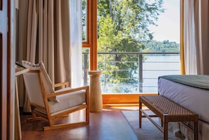 A room with a bed, a wooden chair, a small table, and a view of a lake through large windows. - Puerto Manzano Boutique Hotel (Villa La Angostura)