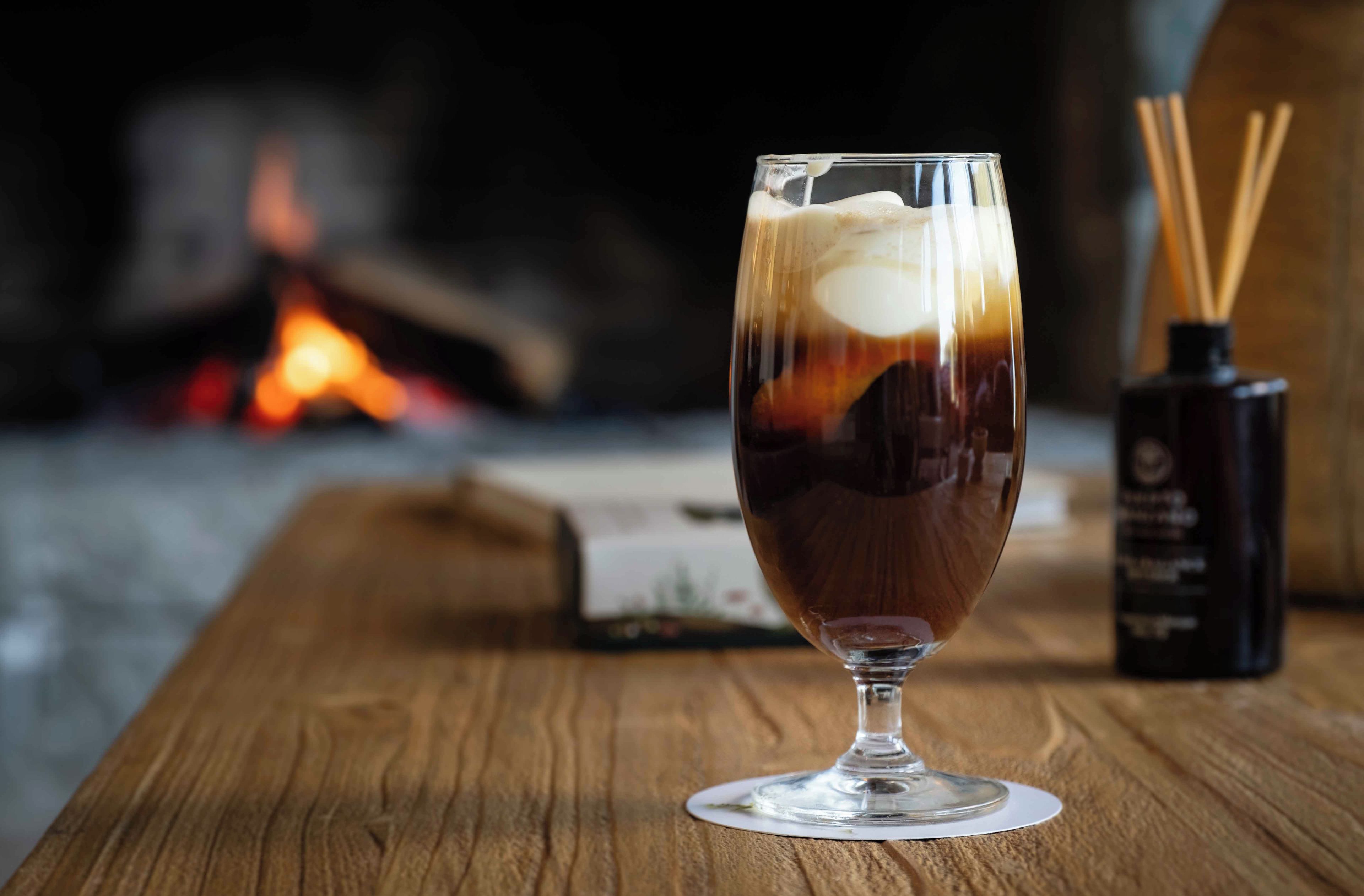 A glass of iced coffee on a wooden table with a candle and reed diffuser in the background.
