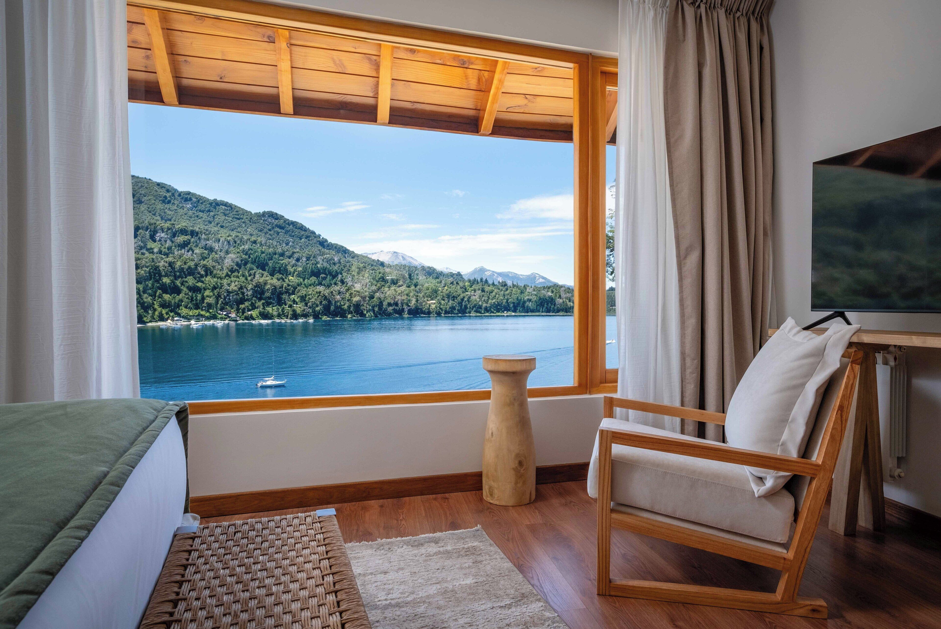 Una habitación con techo de madera, una cama, una silla, una mesita y vistas a un lago y montañas.