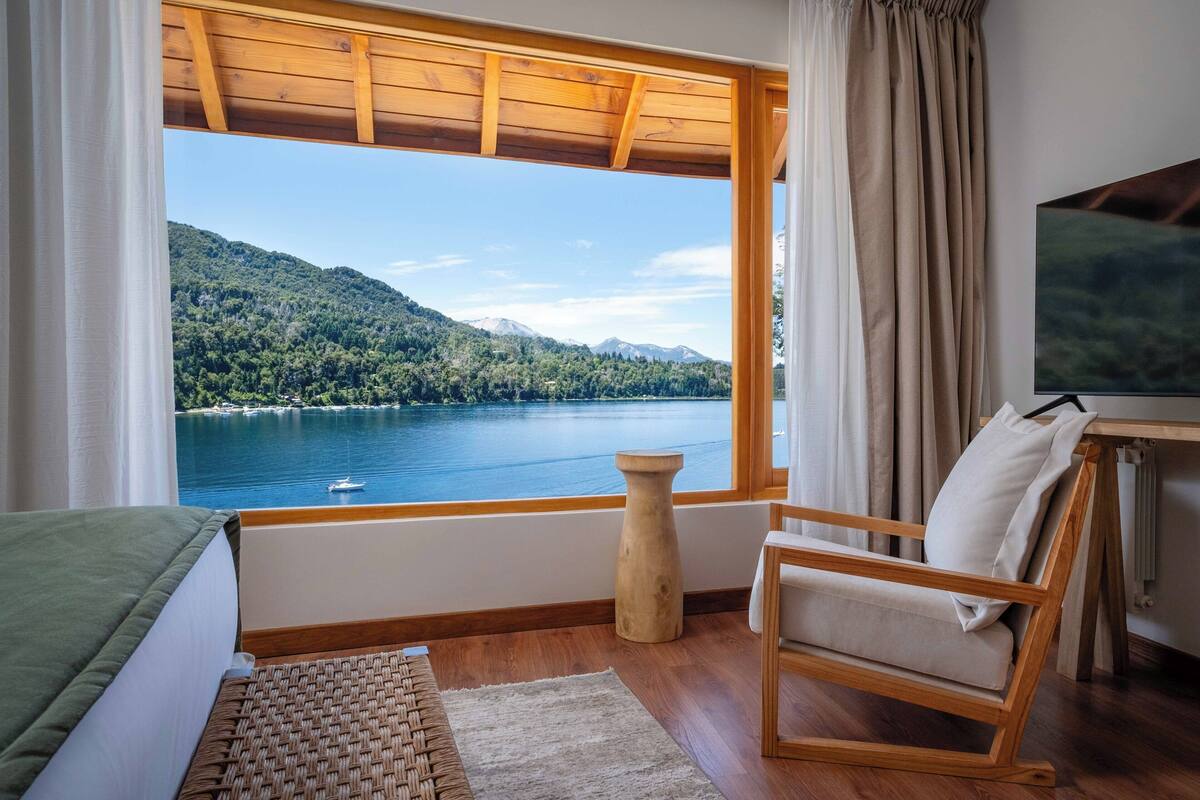 Une pièce avec un plafond en bois, un lit, une chaise, une petite table et une vue sur un lac et des montagnes.