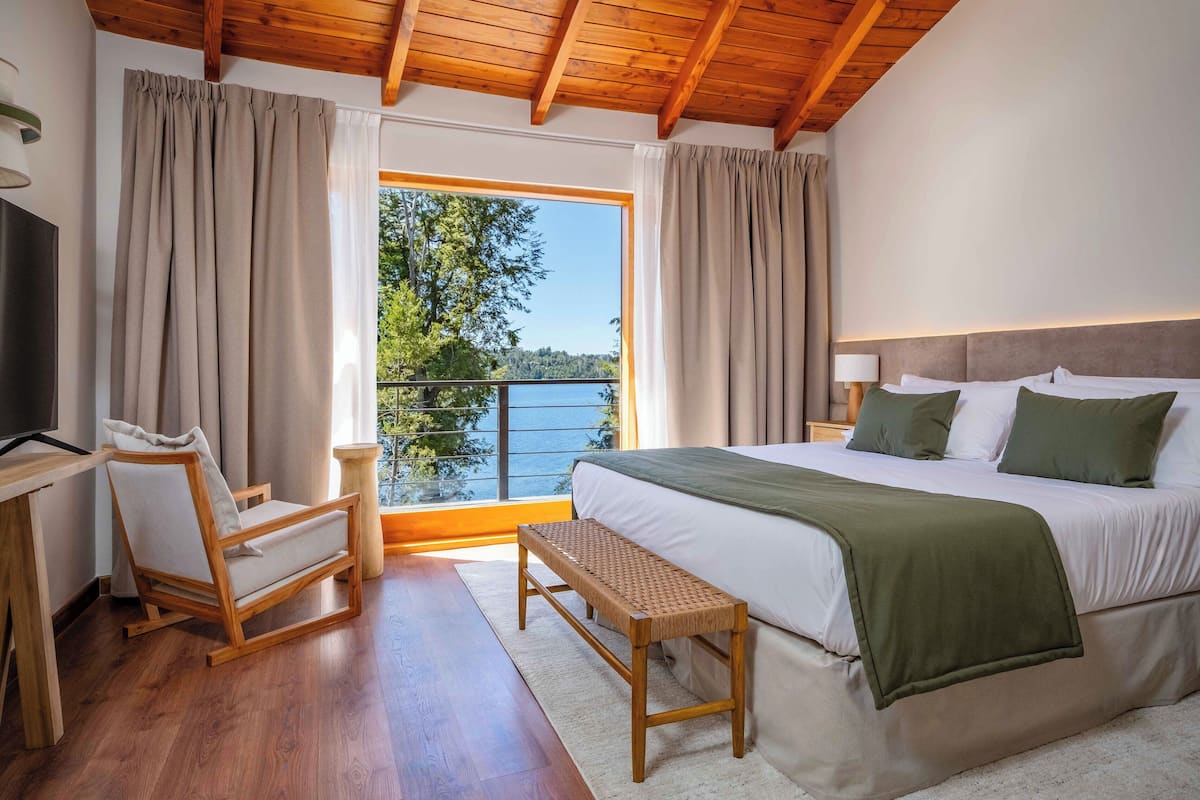 Une chambre à coucher avec un grand lit, une chaise en bois, un banc et une vue sur un lac par la fenêtre.