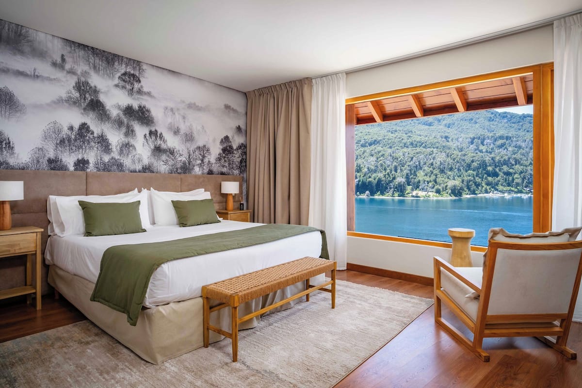 Une chambre à coucher avec un grand lit, un banc, une chaise, une table de chevet et une vue sur un lac et une forêt.