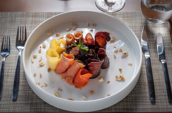 A white plate with various food items including sliced meats and vegetables, accompanied by a fork and knife on a textured tablecloth. - Puerto Manzano Boutique Hotel (Villa La Angostura)