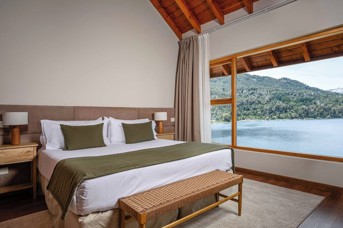 Une chambre avec une grande fenêtre, un lit avec un oreiller vert, un banc en bois et une table de chevet avec une lampe.