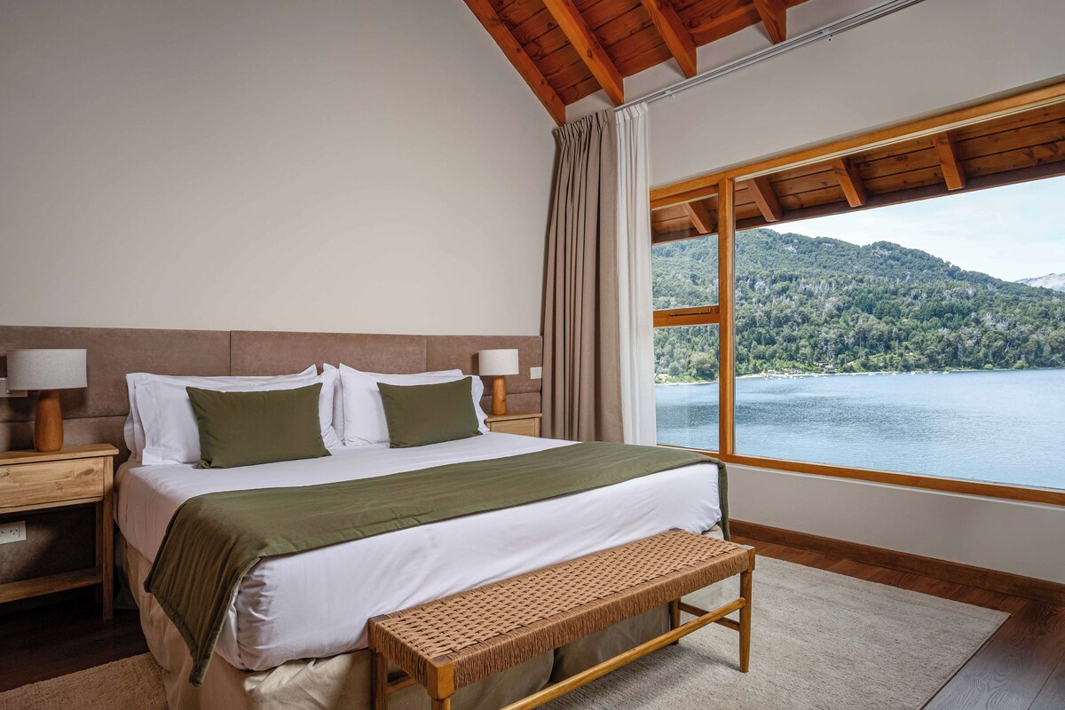 A bedroom with a large window, a bed with a green pillow, a wooden bench, and a nightstand with a lamp.