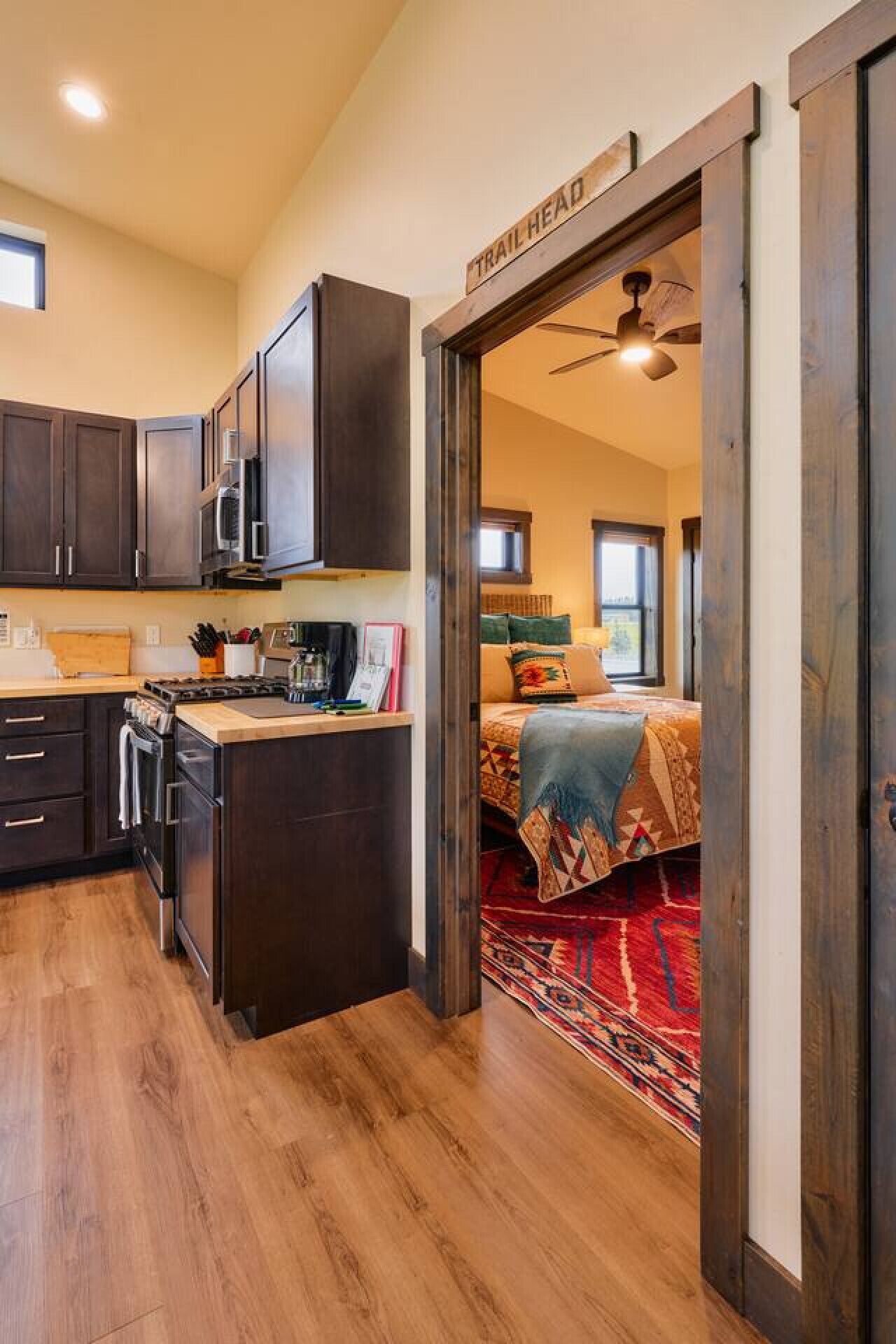 A modern kitchen with dark wood cabinets, a stove, and a microwave. There is a wooden island with a sink and a window above it. The kitchen leads to a bedroom with a bed, a window, and a red patterned rug.
