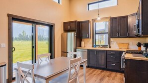 A modern kitchen with a dining area, large windows, and a view of a green landscape.