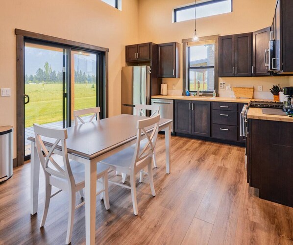 A modern kitchen with a dining area, large windows, and a view of a green landscape. - Teakettle - Stunning Mountain Views, Close to GNP! (Columbia Falls)