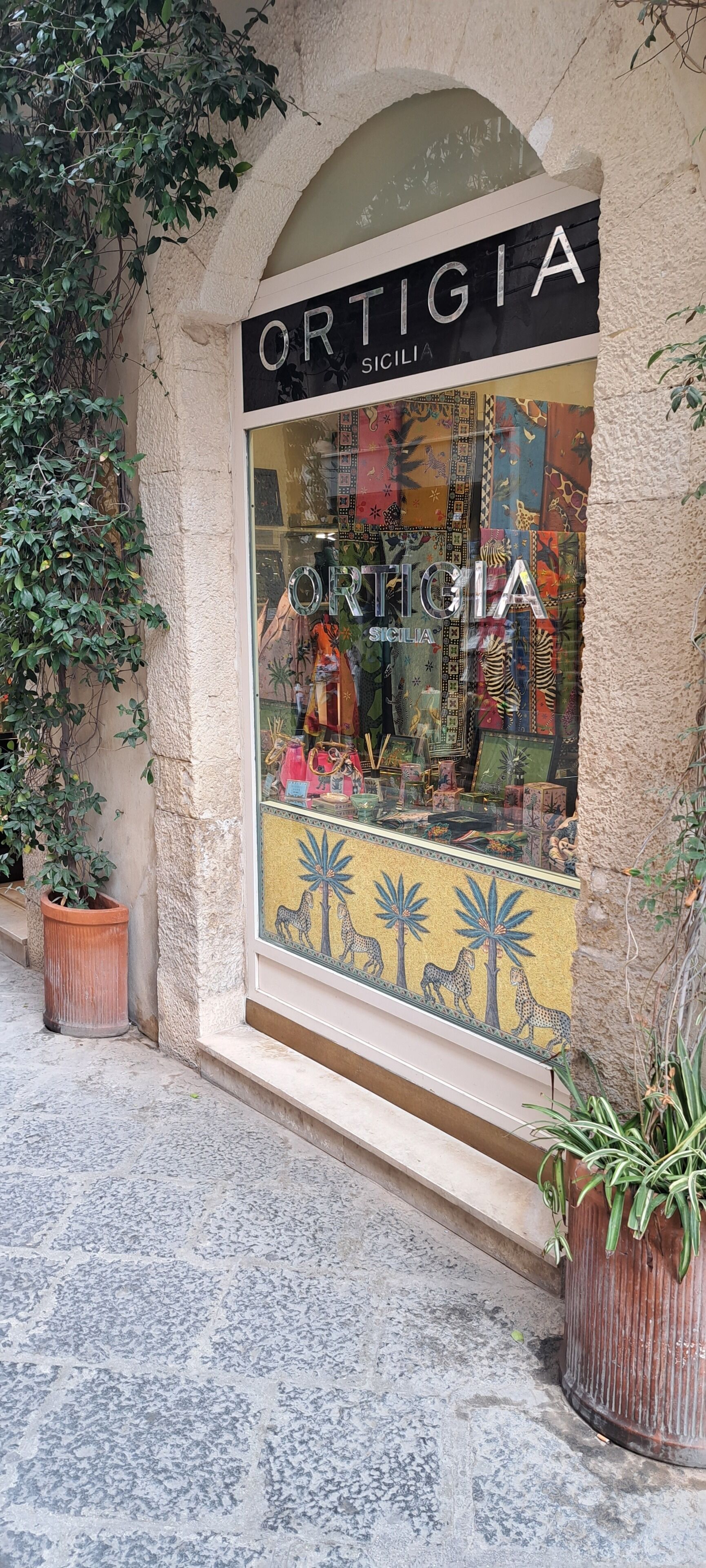 En butiksfasad med en skylt som visar texten ”ORTIGIA SICILIA” och ett skyltfönster med mönstrat tyg och en mosaik.