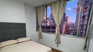 Een slaapkamer met een bed, gordijnen, een raam en uitzicht op gebouwen.