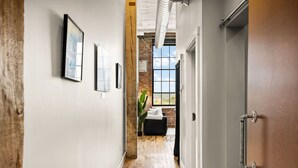 Un couloir avec un plancher en bois, des murs blancs et des tableaux encadrés. Il y a une porte sur la droite et une fenêtre au fond du couloir.