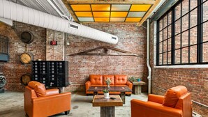 Une pièce aux murs en briques, avec une grande fenêtre, des canapés en cuir orange, une table basse en bois et un meuble de rangement noir.
