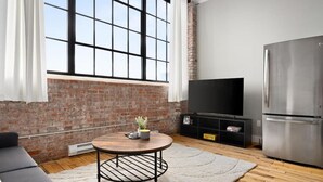 A modern living room with a brick wall, a large window, a round coffee table, a black sofa, a flat-screen TV, and a stainless steel refrigerator.